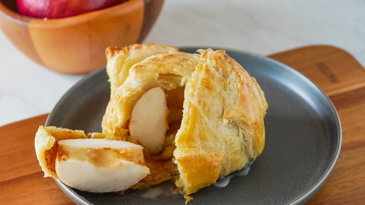 A baked apple wrapped in flaky pastry sits on a gray plate with one slice cut.