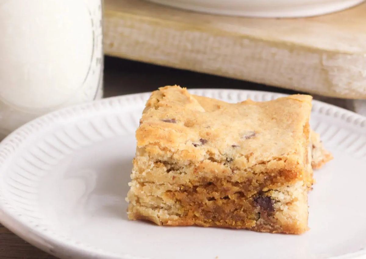 A square piece of cookie bar with chocolate chips sits on a white plate, with a glass of milk in the background.