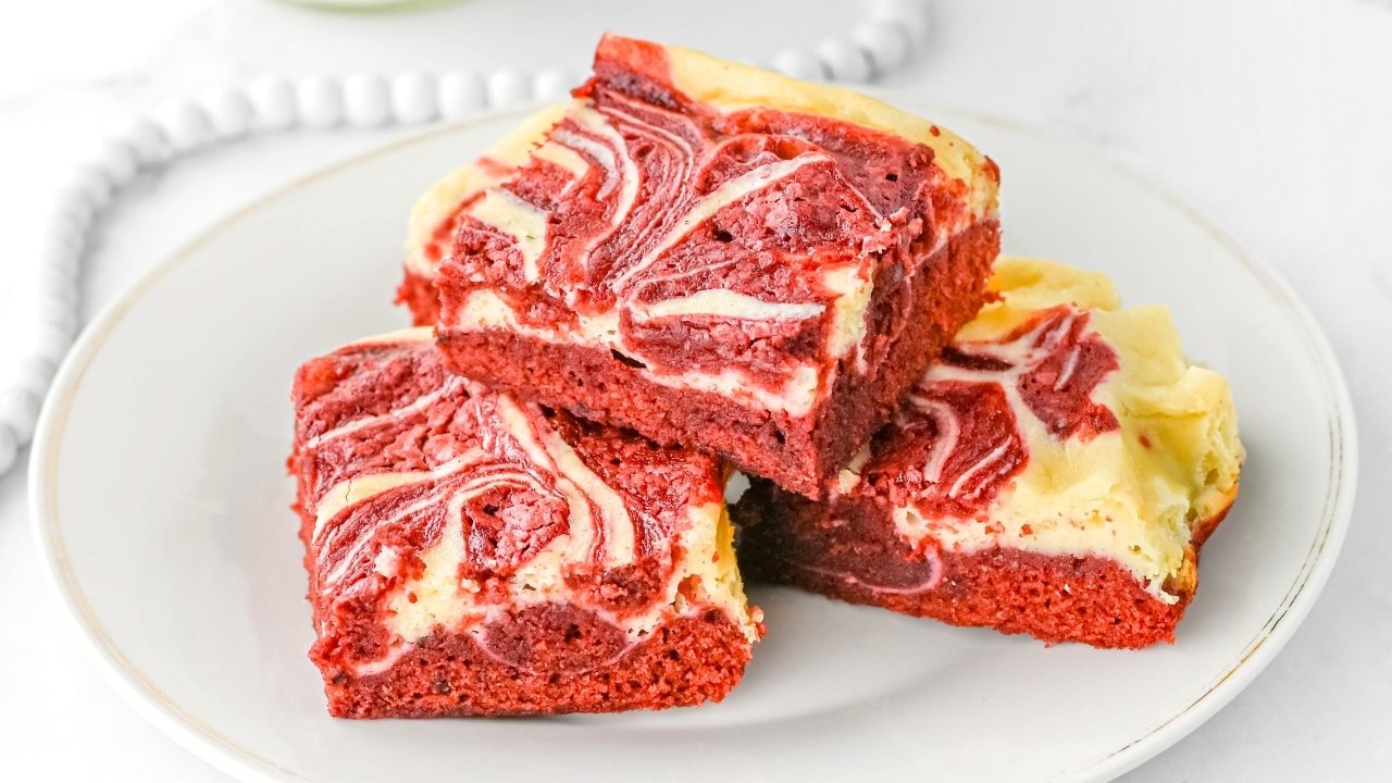 Three red velvet cheesecake brownies with a marbled cream cheese swirl are stacked on a white plate.