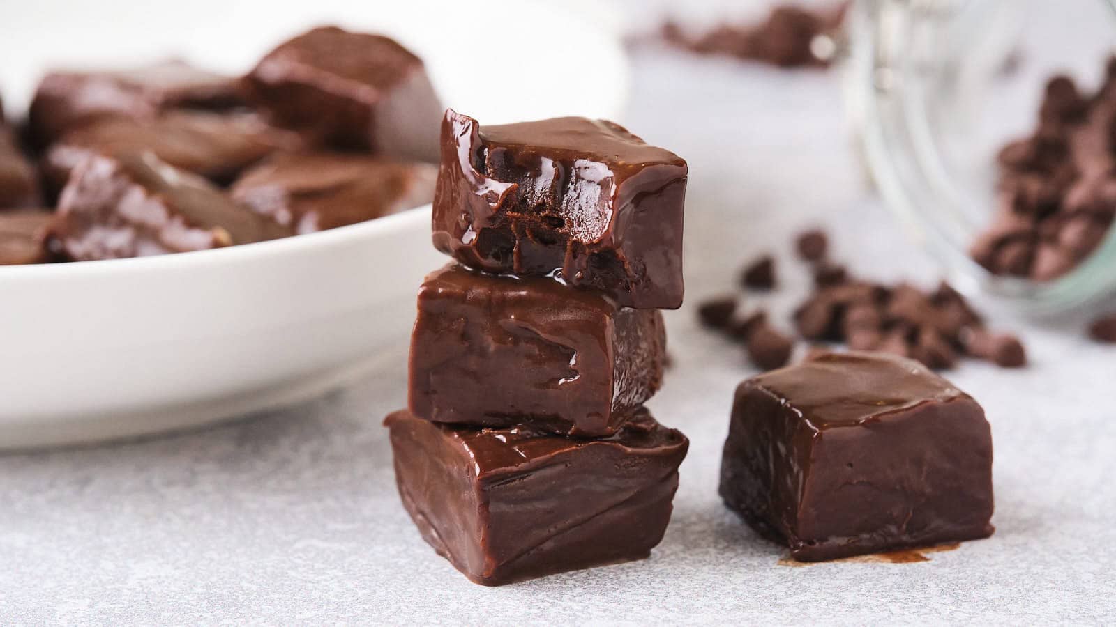 Four glossy, square-shaped pieces of chocolate fudge stacked on a light surface, with a plate of more fudge and scattered chocolate chips in the background.