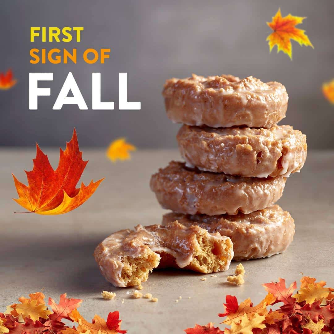 Four glazed donuts are stacked on a surface with one partially eaten in front; autumn leaves and the text First Sign of Fall are visible.