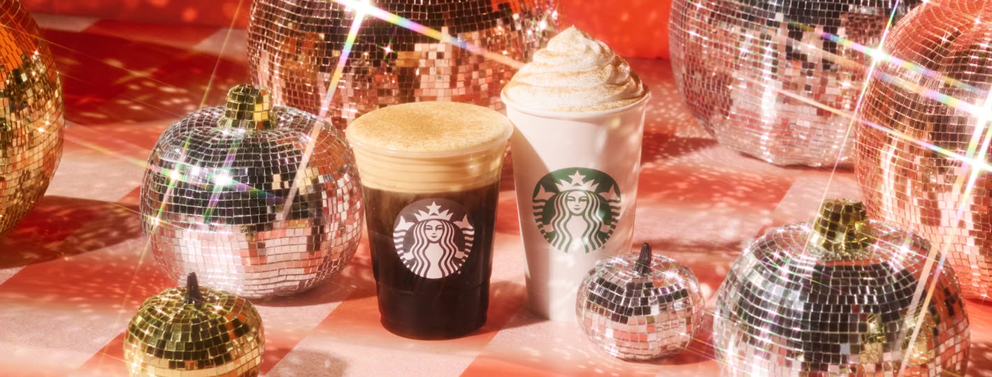 Two Starbucks drinks, one iced and one hot, are placed among several disco ball pumpkins on a red and white checkered surface with sparkling light reflections.
