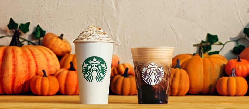 Two Starbucks drinks, one hot with whipped cream and one iced, are placed on a wooden surface with several pumpkins in the background.