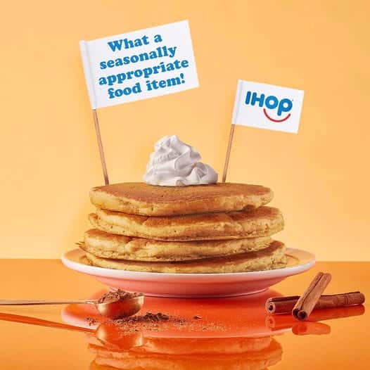 A stack of pancakes topped with whipped cream, with two mini flags—one reading What a seasonally appropriate food item! and the other displaying the IHOP logo. Cinnamon sticks and powder nearby.