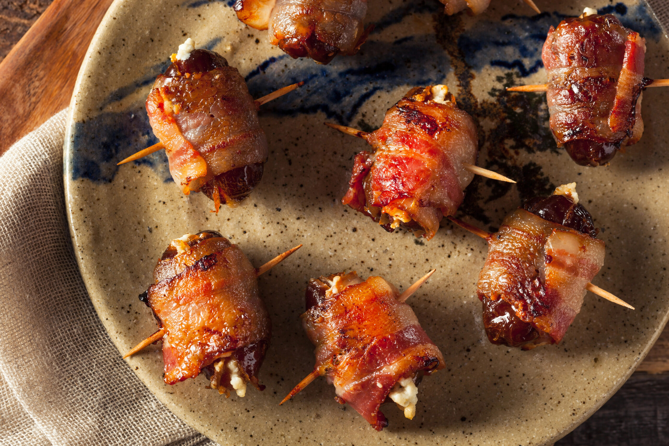 A ceramic plate with bacon-wrapped dates secured by toothpicks, arranged on a textured surface.