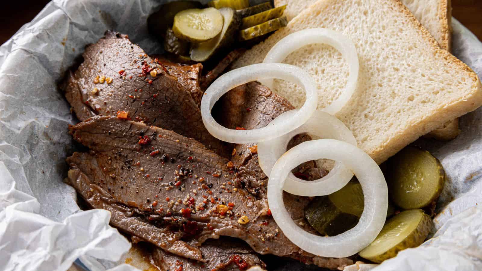 Slices of seasoned brisket topped with onion rings are served alongside sliced pickles and two pieces of white bread on a sheet of parchment paper.