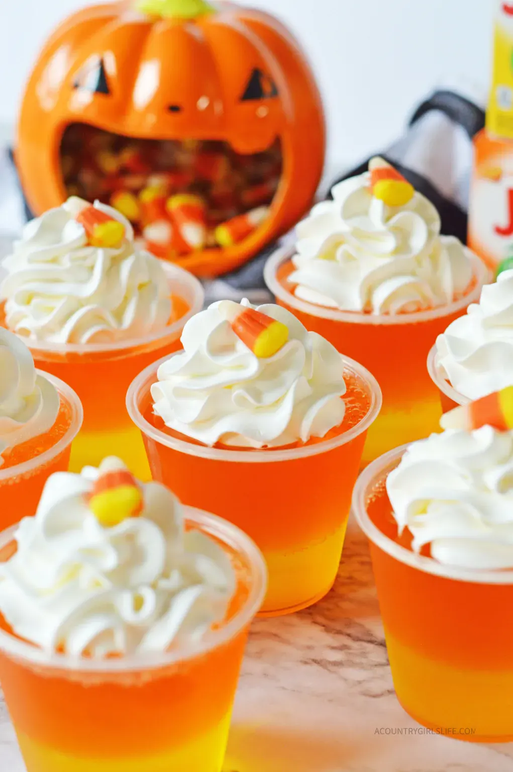 Several orange and yellow layered gelatin cups topped with whipped cream and candy corn, with a pumpkin-shaped container of candy corn in the background.