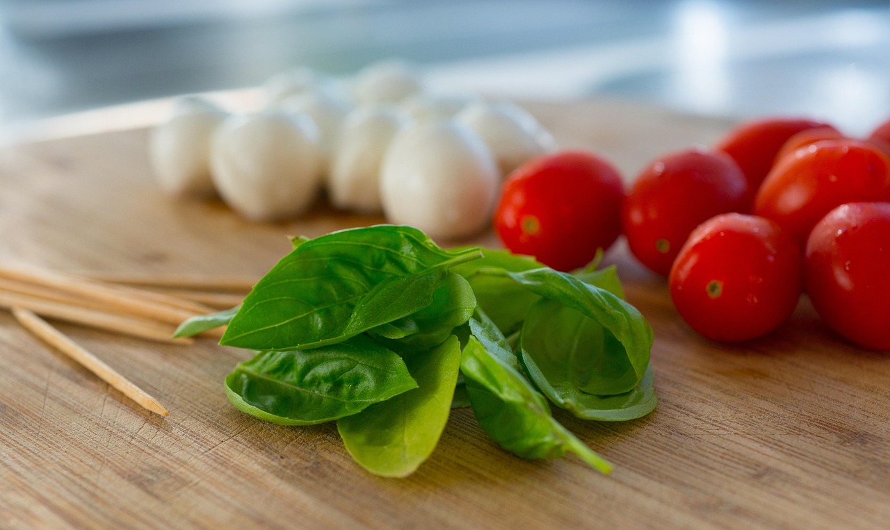 Fresh basil leaves, cherry tomatoes, and mozzarella balls arranged on a wooden cutting board with wooden skewers nearby.
