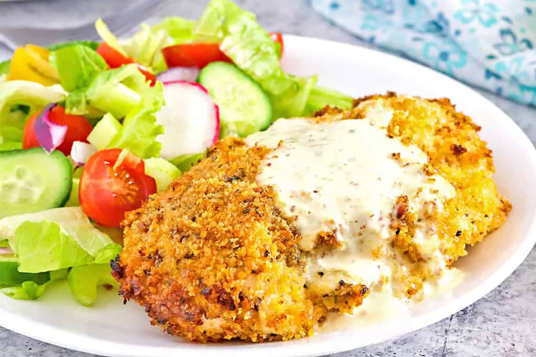 A plate with breaded chicken topped with creamy sauce, served alongside a fresh salad with lettuce, cucumbers, tomatoes, and radishes.