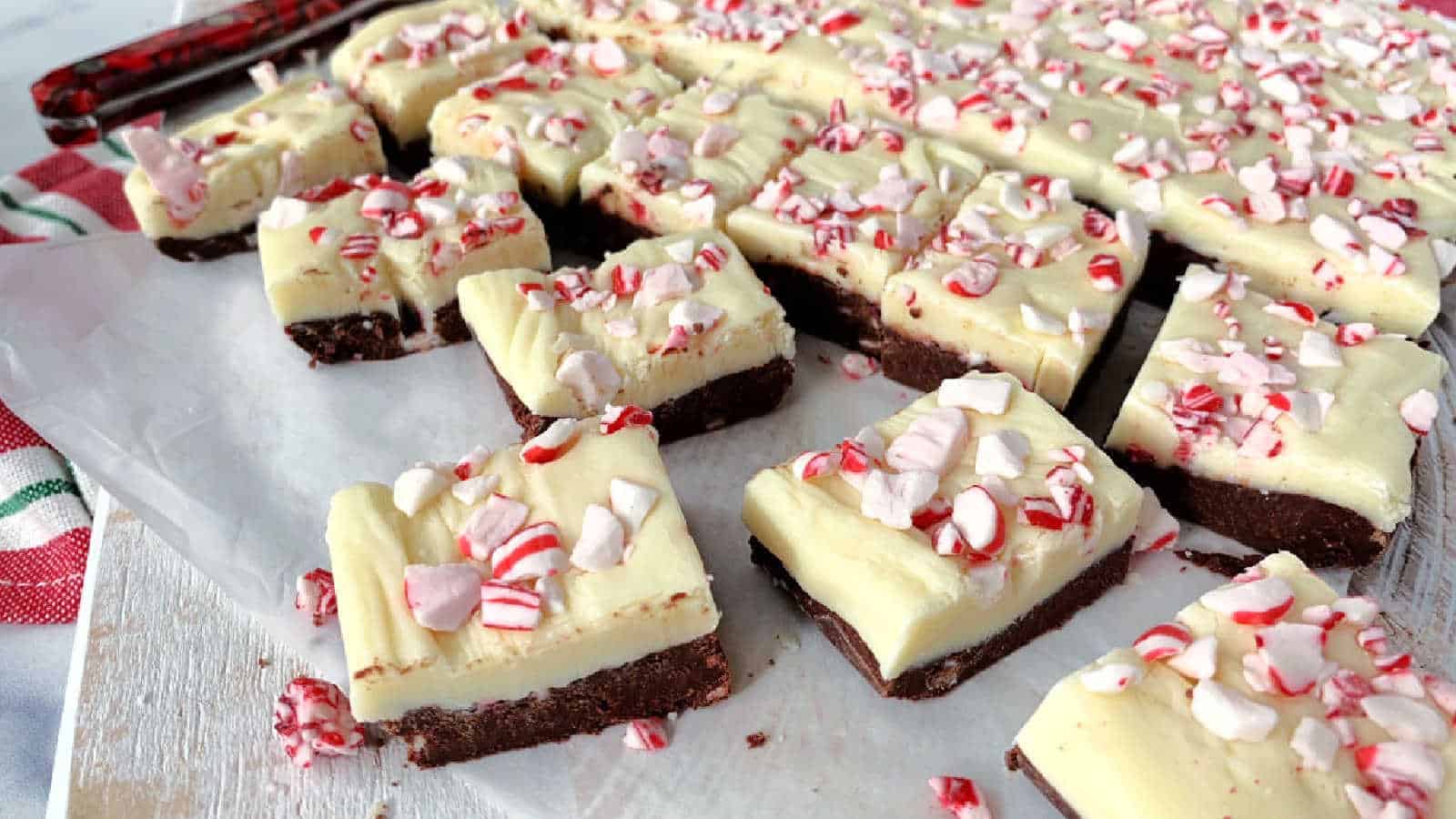 Rectangular pieces of layered fudge with a chocolate base, white topping, and crushed peppermint candy sprinkled on top, arranged on parchment paper.