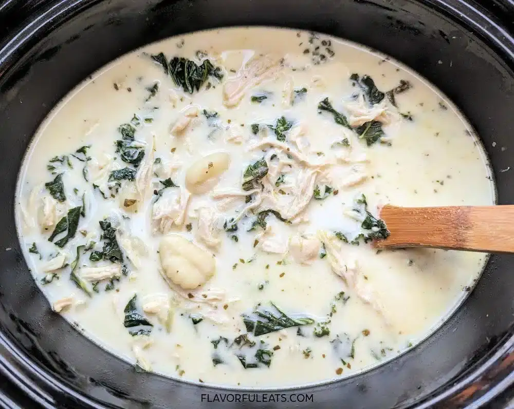 A creamy soup with shredded chicken, gnocchi, and spinach in a slow cooker, being stirred with a wooden spoon.