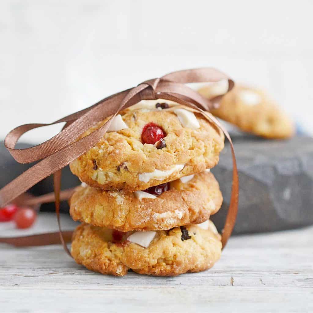 A stack of three cookies with visible nuts and berries, tied together with a brown ribbon, placed on a light wooden surface.