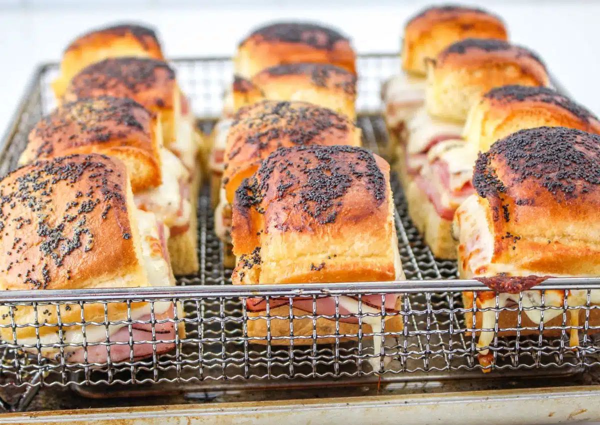 A tray of baked ham and cheese sliders in a wire rack, topped with poppy seeds and arranged in rows.