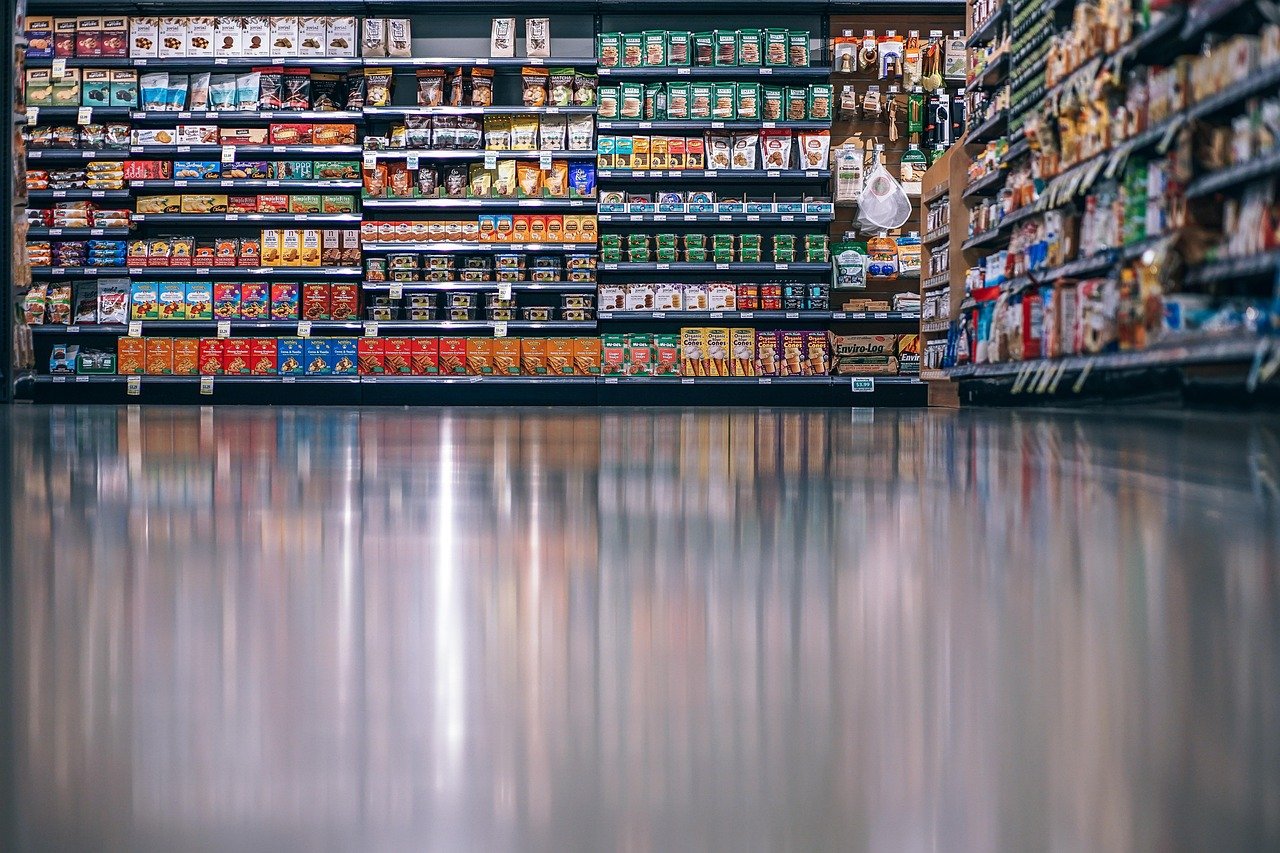 A supermarket aisle with shelves stocked with various packaged food products and a shiny, empty floor in the foreground.