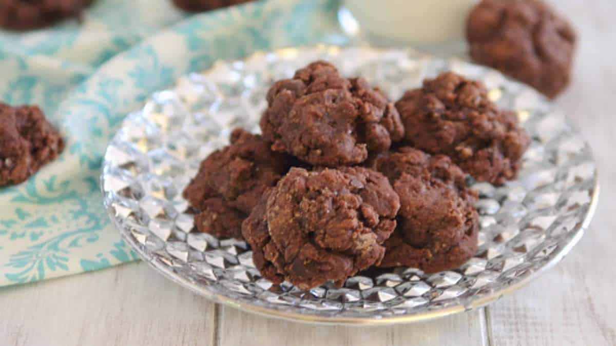 A clear glass plate holds several chocolate cookies on a light wooden surface, with part of a patterned napkin visible in the background.