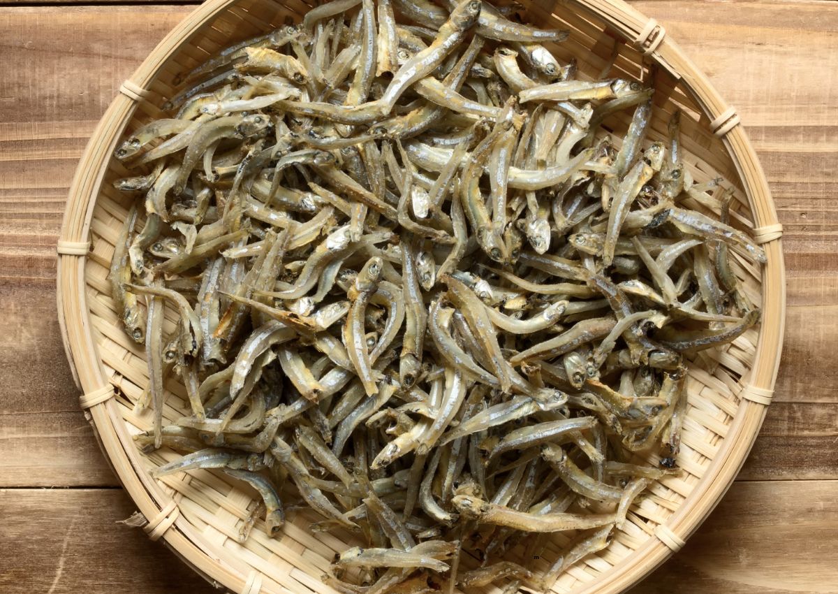 A round bamboo basket filled with dried small fish, placed on a wooden surface.