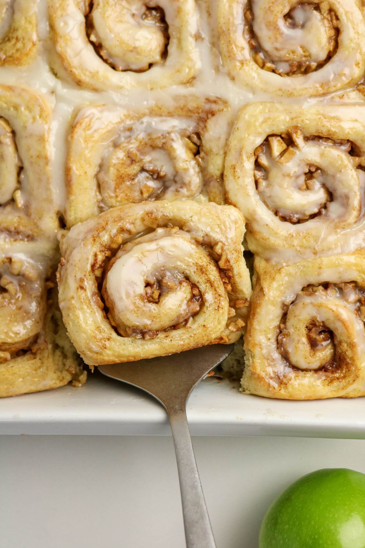 A metal spatula lifts a glazed cinnamon roll from a tray of cinnamon rolls; a green apple is visible in the bottom right corner.