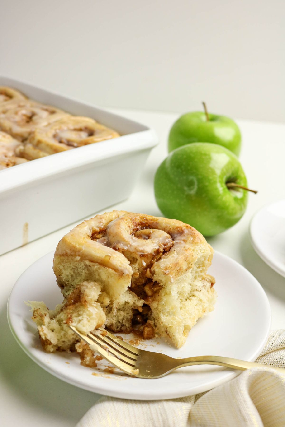 A cinnamon roll with icing is served on a plate with a fork, two green apples, and a baking dish of more rolls in the background.
