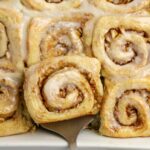 Close-up of glazed cinnamon rolls with chopped nuts in a baking dish, one roll being lifted with a spatula.