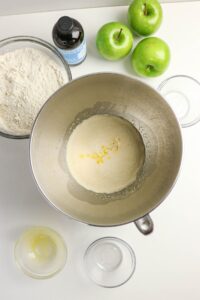 A mixing bowl with wet ingredients, surrounded by bowls of flour, eggs, vanilla extract, and three green apples on a white surface.