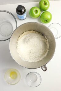 A mixing bowl with flour, surrounded by three green apples, a bottle of vanilla extract, and several empty glass bowls on a white surface.