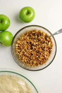Chopped green apples mixed with cinnamon in a glass bowl, three whole green apples beside it, and covered dough in another bowl on a white surface.