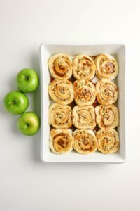 A rectangular white baking dish with twelve unbaked cinnamon rolls next to three green apples on a white surface.