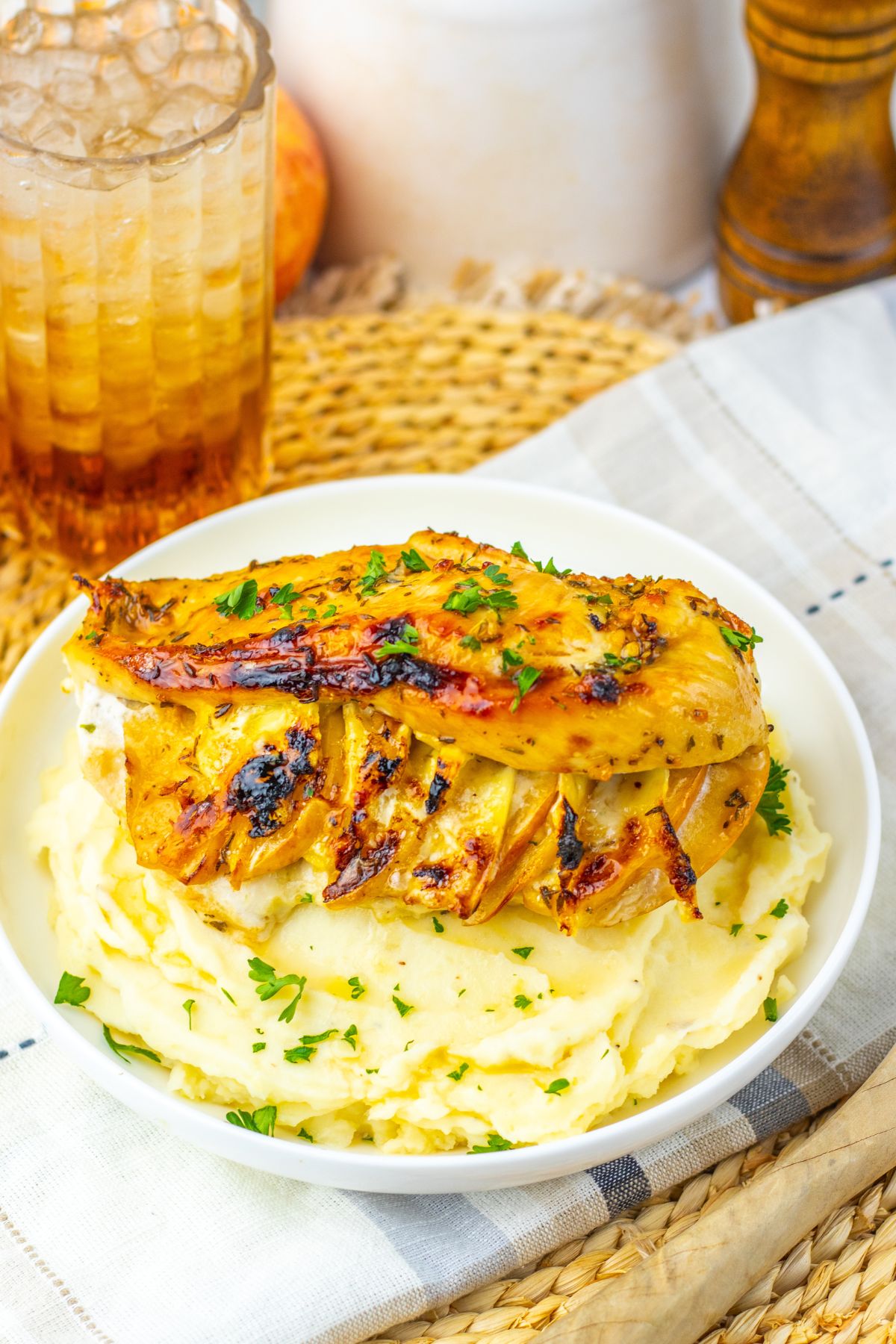 Grilled chicken breast garnished with herbs served on a bed of mashed potatoes in a white bowl, with a glass of iced drink in the background.