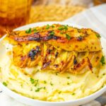 Grilled chicken breast stuffed with cheese sits on a mound of mashed potatoes, garnished with chopped parsley.