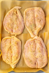 Four raw, seasoned chicken breasts placed on a parchment-lined baking tray.