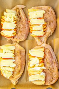 Four raw chicken breasts, each sliced open and stuffed with alternating slices of apple and brie cheese, are arranged on parchment paper and seasoned with pepper.