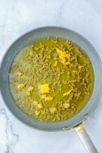 A frying pan with oil, cumin seeds, and chunks of yellow turmeric or ginger on a marble surface.