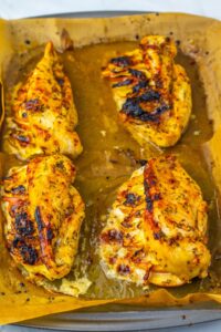 Four seasoned and baked chicken breasts on parchment paper in a baking tray, with visible browned and slightly charred spots.