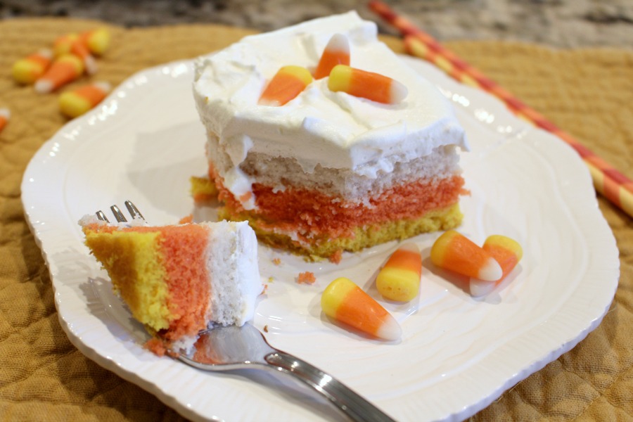 A slice of layered cake with white frosting and candy corn on top, served on a white plate with a fork and extra candy corn pieces.