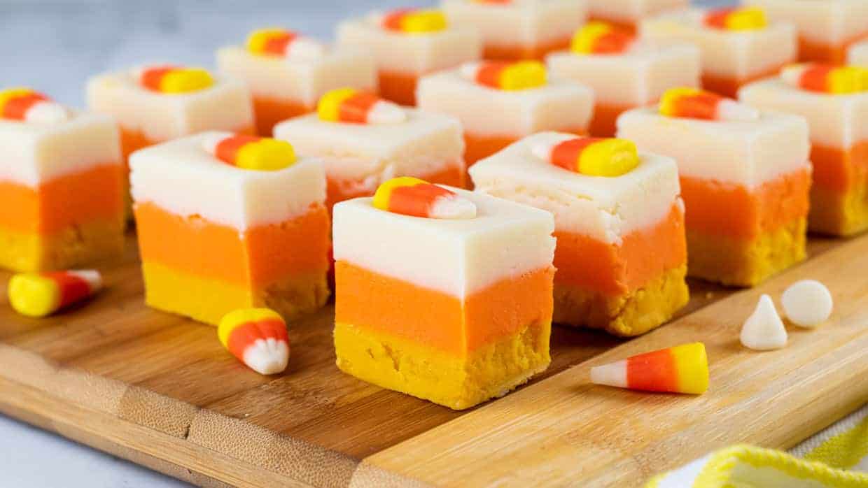 Rows of square, layered fudge pieces in white, orange, and yellow colors, each topped with a candy corn, displayed on a wooden board.