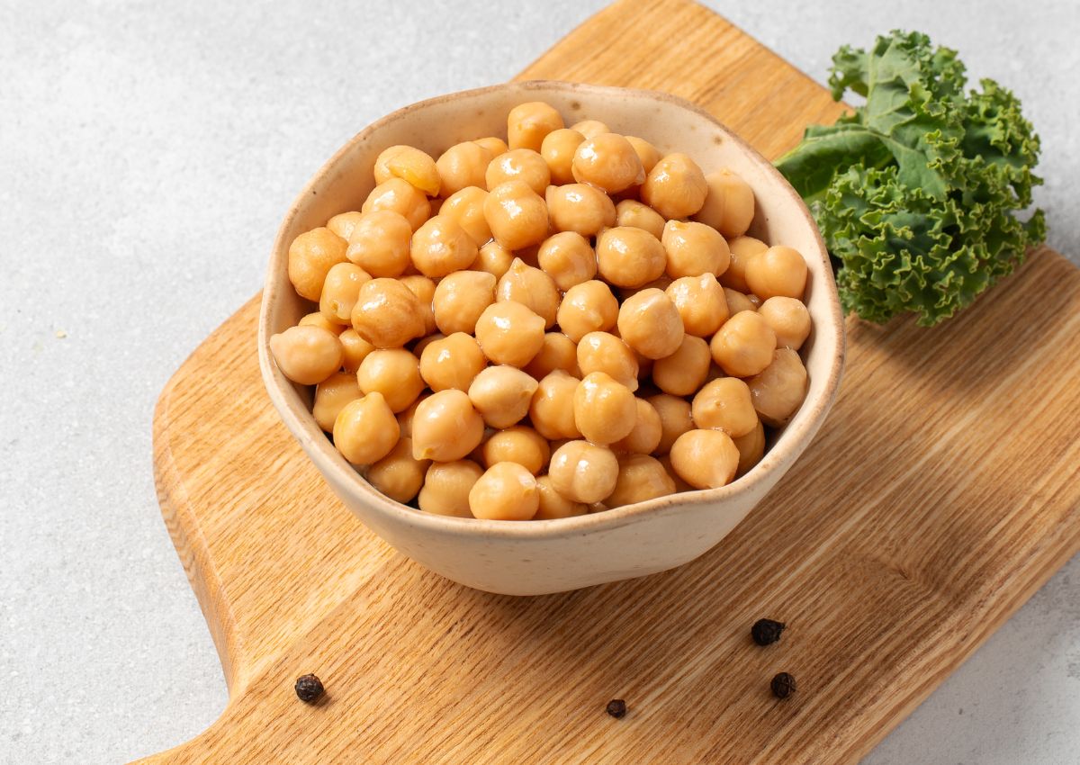A bowl of cooked chickpeas on a wooden board, with a piece of kale and peppercorns nearby.