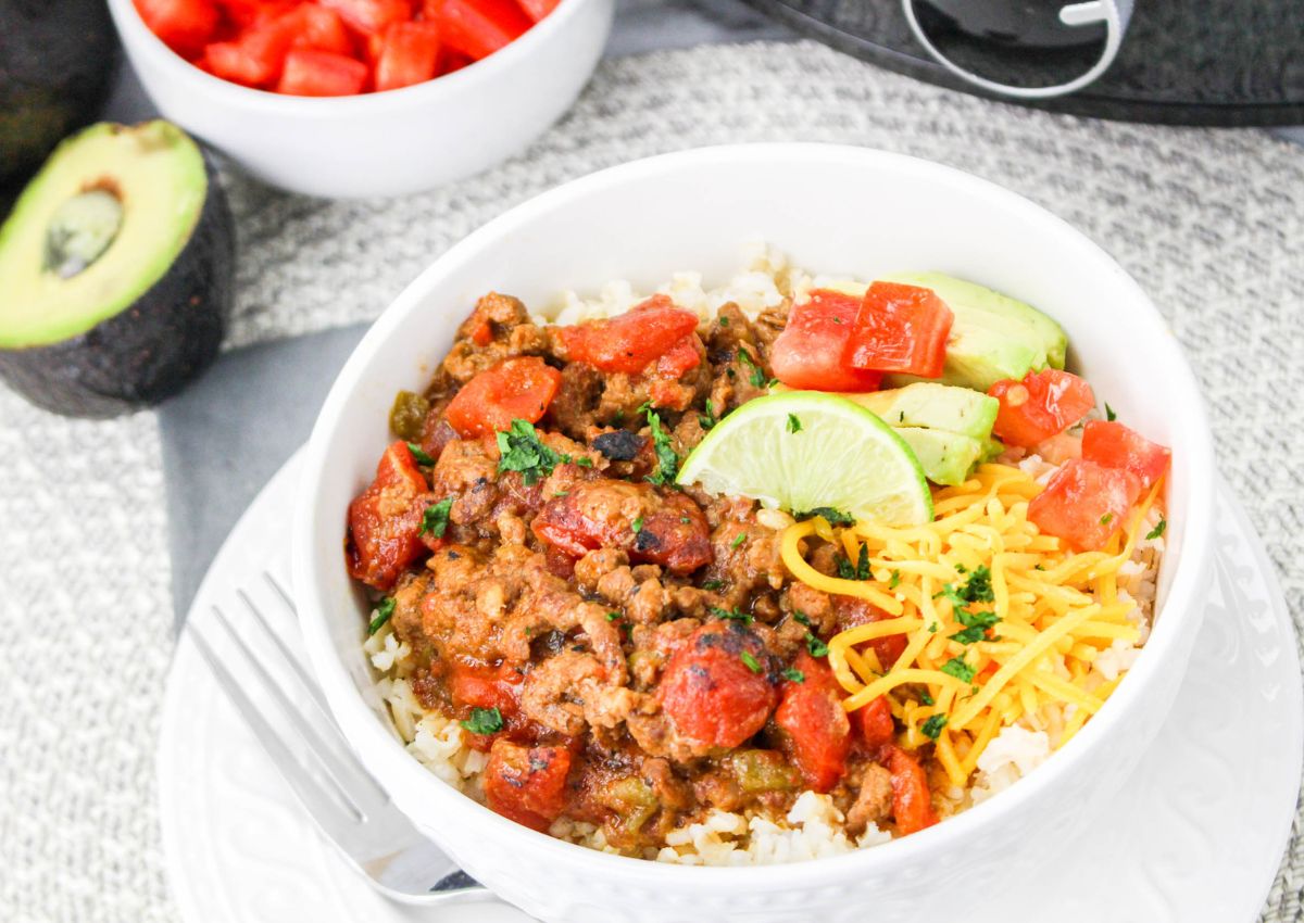 A bowl of rice topped with seasoned ground beef, kidney beans, diced tomatoes, shredded cheese, avocado slices, lime wedges, and chopped herbs. A fork is placed in the bowl.