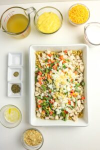 Ingredients for a casserole are arranged around a dish containing chopped chicken, mixed vegetables, and onions on top of noodles. Separate bowls hold soup, cheese, broth, milk, spices, butter, and crumbs.
