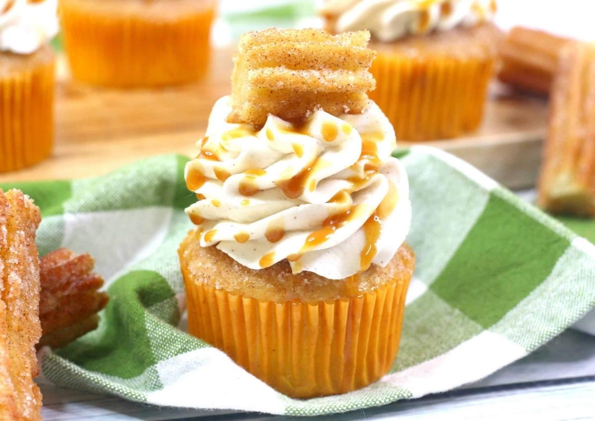 Cupcake with swirled frosting, caramel drizzle, and churro pieces on top, set on a green and white checkered cloth.