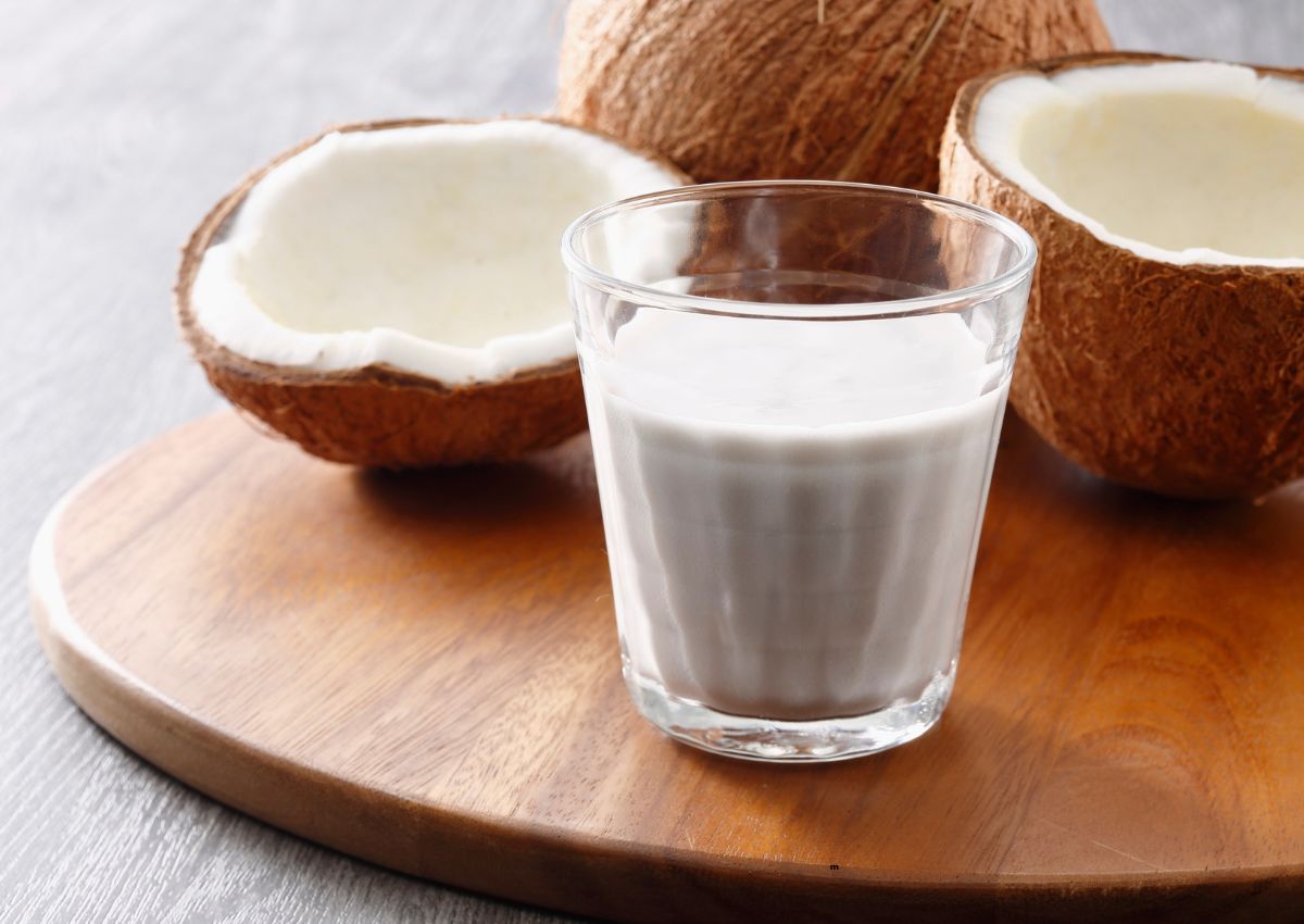 A glass of coconut milk sits on a wooden board next to halved coconuts.