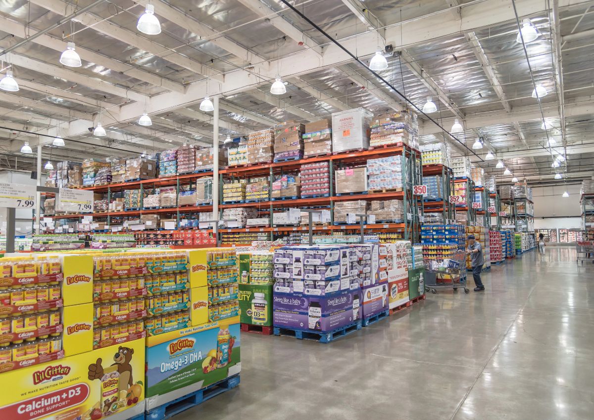 Wide view of a warehouse-style grocery store with high shelves stocked with bulk items; a shopper pushes a cart down a spacious aisle.