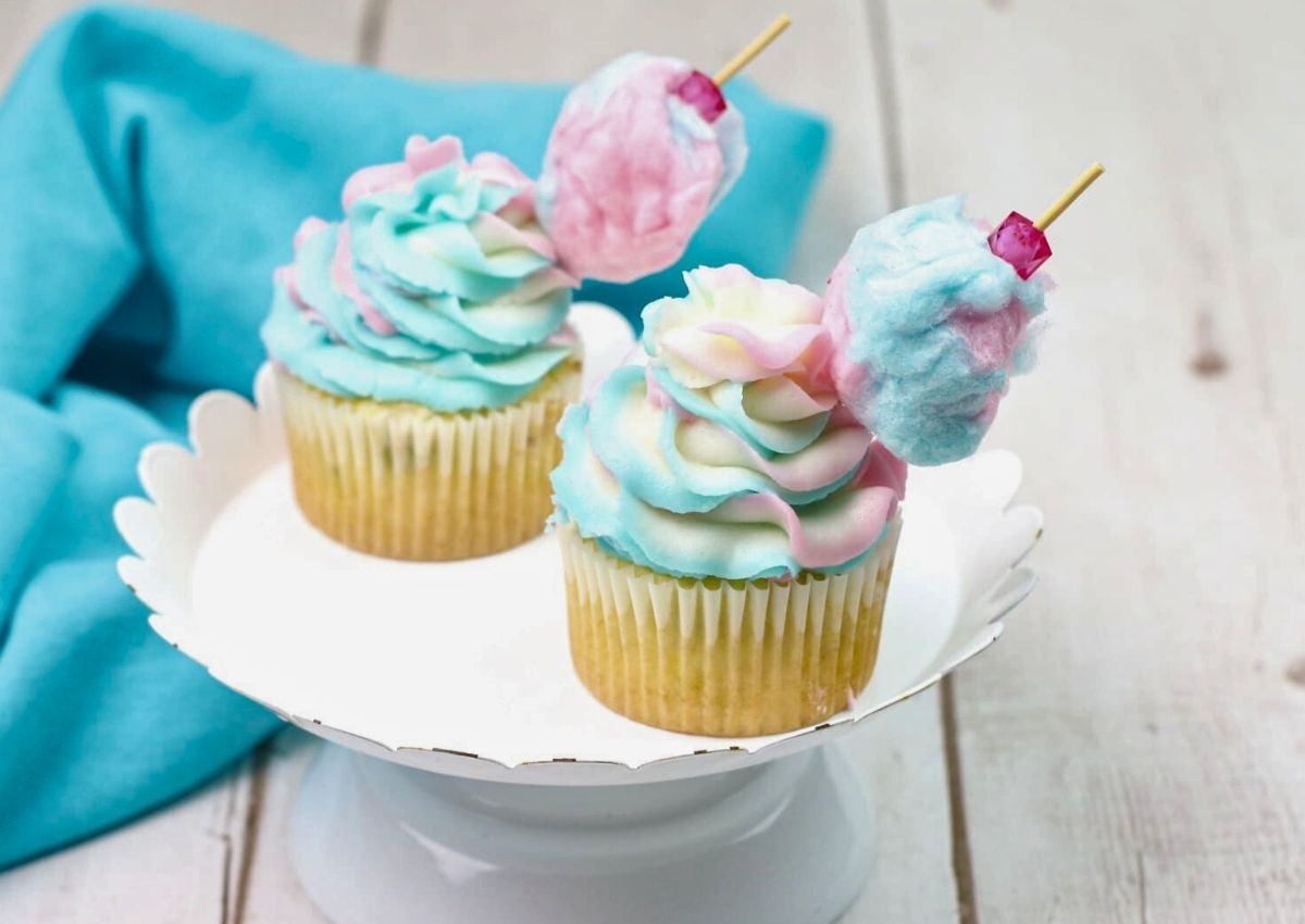 Two cupcakes with pastel swirled frosting and cotton candy decorations on top, displayed on a white cake stand with a blue cloth in the background.