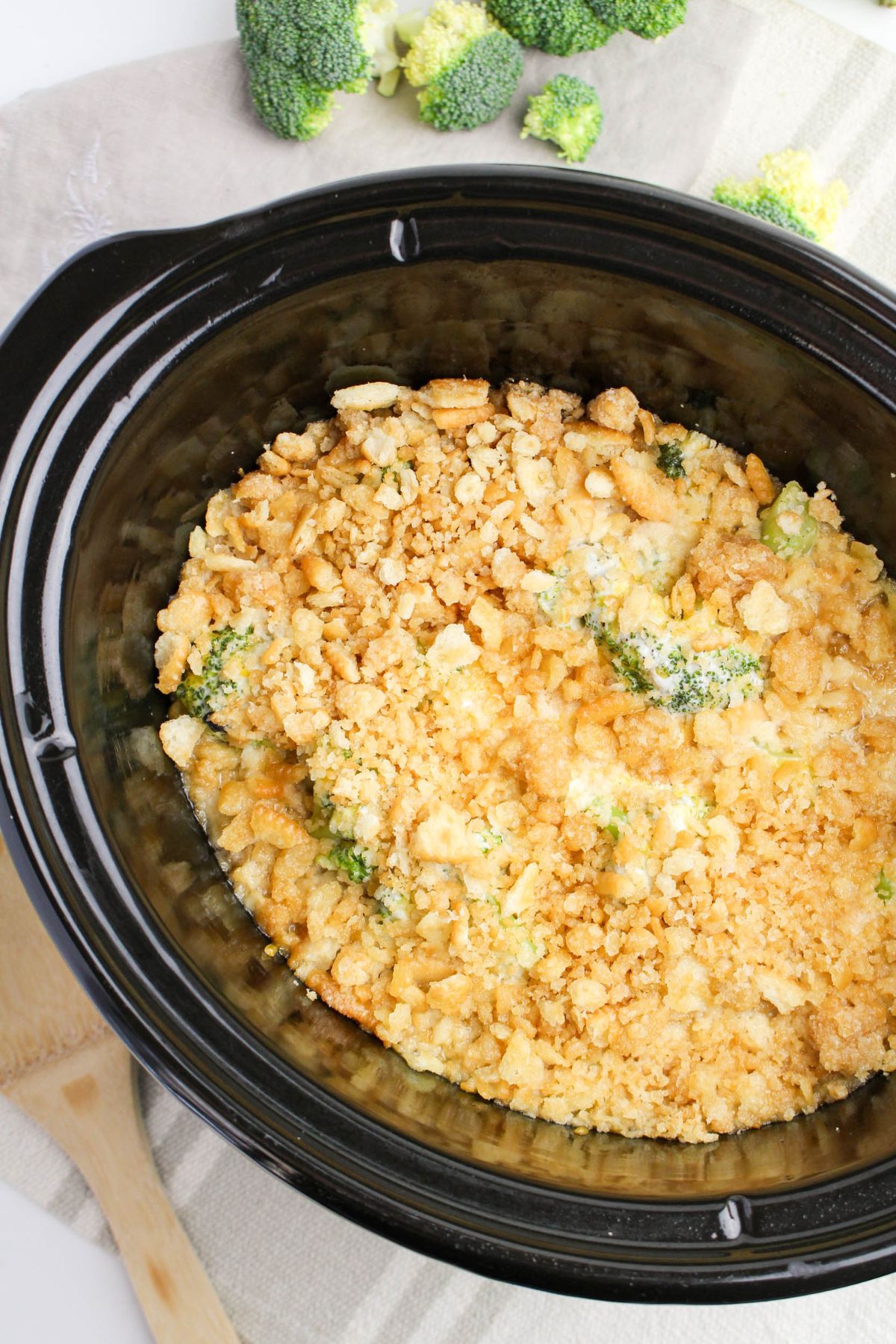 A slow cooker filled with broccoli casserole topped with a golden breadcrumb crust, sitting on a beige napkin with raw broccoli florets nearby.