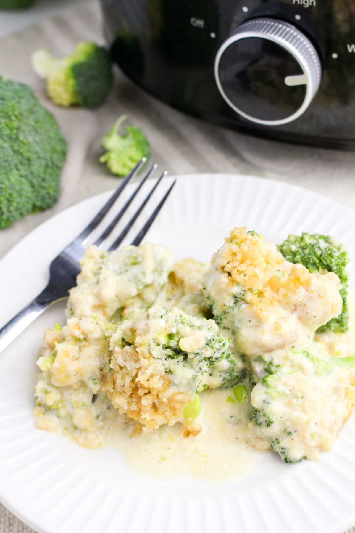A plate of creamy broccoli casserole with a fork beside it, set in front of a slow cooker and broccoli florets.