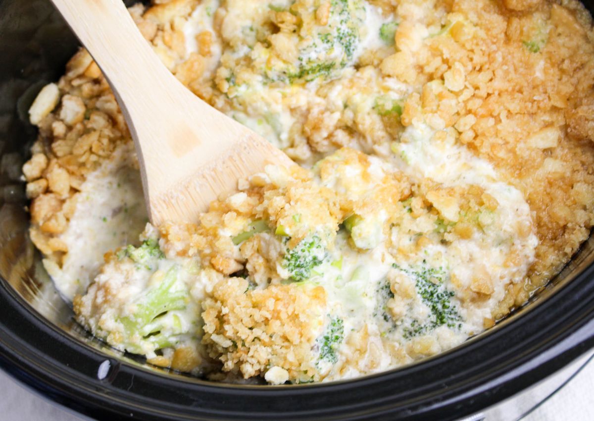 A close-up of a broccoli casserole with a creamy sauce and crunchy topping, being stirred with a wooden spoon inside a slow cooker.