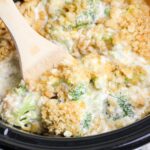 A wooden spoon scoops creamy broccoli casserole topped with a golden, crumbly crust inside a black dish.
