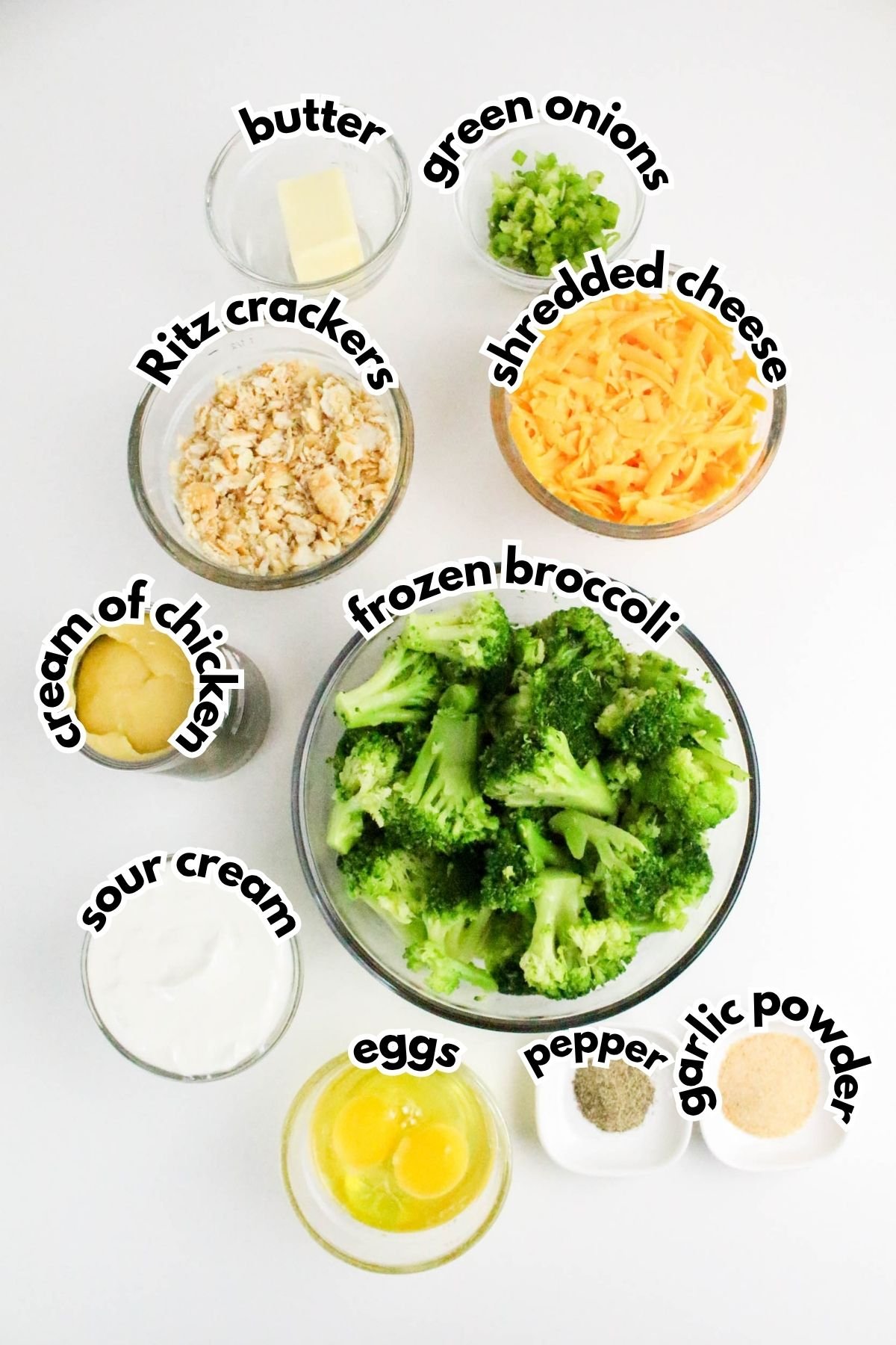 Bowls containing frozen broccoli, shredded cheese, green onions, butter, Ritz crackers, cream of chicken, sour cream, eggs, pepper, and garlic powder are arranged on a white surface.