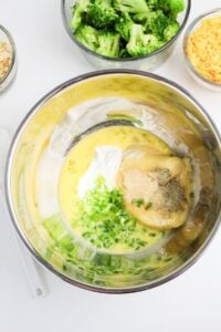 A metal bowl containing eggs, sour cream, chopped green onions, seasonings, and soup mixture, with bowls of broccoli, cheese, and fried onions nearby.
