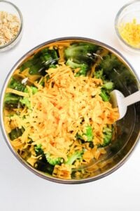 A mixing bowl filled with broccoli florets and shredded cheddar cheese, with a white spatula inside and two small bowls nearby.
