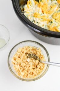 A glass bowl with a crumbly mixture and a spoon sits next to a slow cooker containing a cheese-topped dish. A smaller bowl with liquid is also visible.